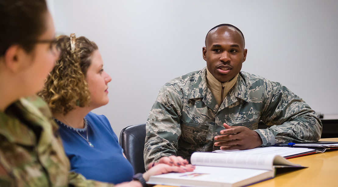 Male student in military fatigues sitting at desk with two female students – one in military fatigues, in discussion. 