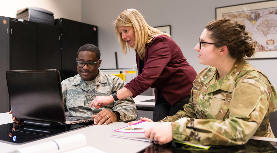 A female teacher points to a laptop screen while talking to a male student as a female student (both in fatigues) listens on. 