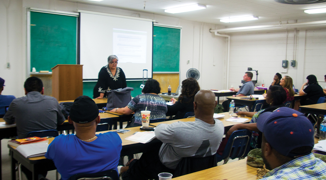 A female teacher standing in front of a blackboard at the head of a classroom talking to a dozen adult men and women students seated at desks. 