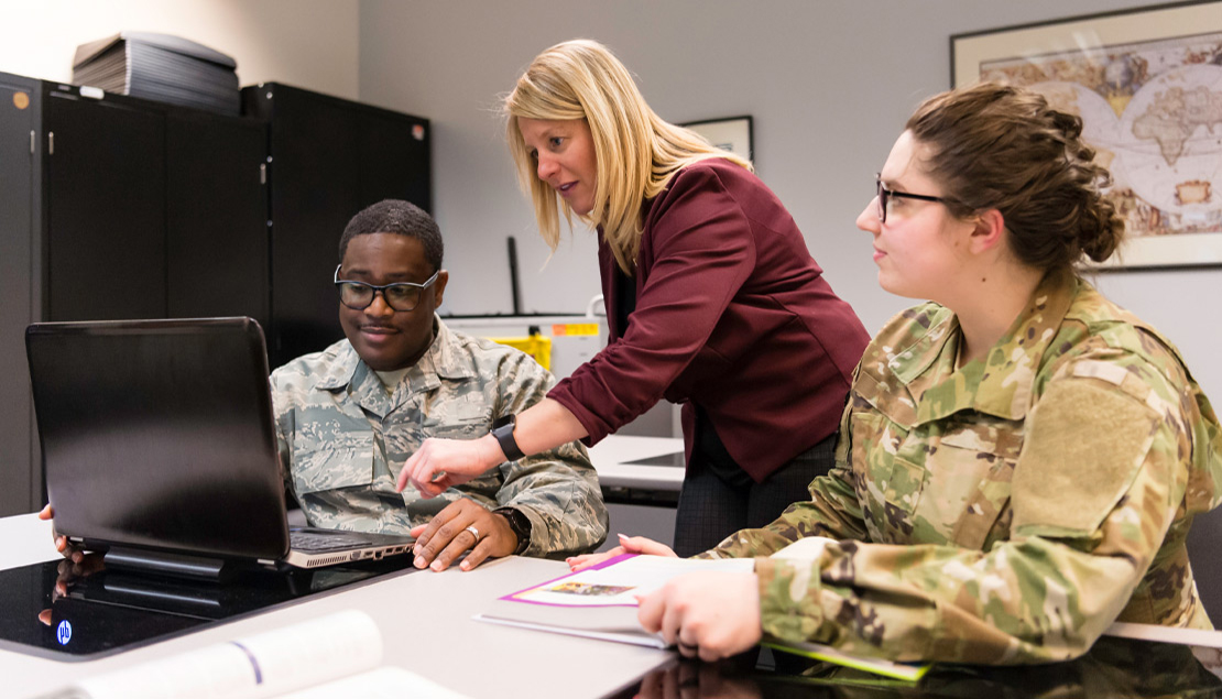 A teacher assists two students in two different branches of military fatigues in a computer classroom situation. 