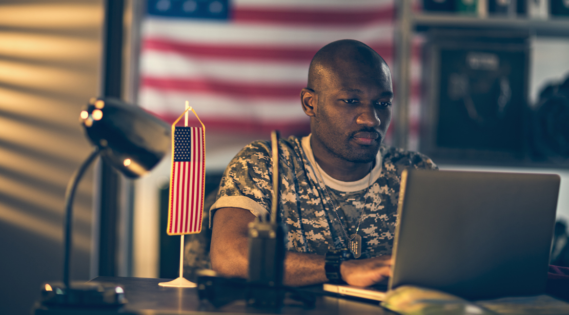 Male student in military fatigues working on a laptop for an online class with an American flag behind him on the wall.