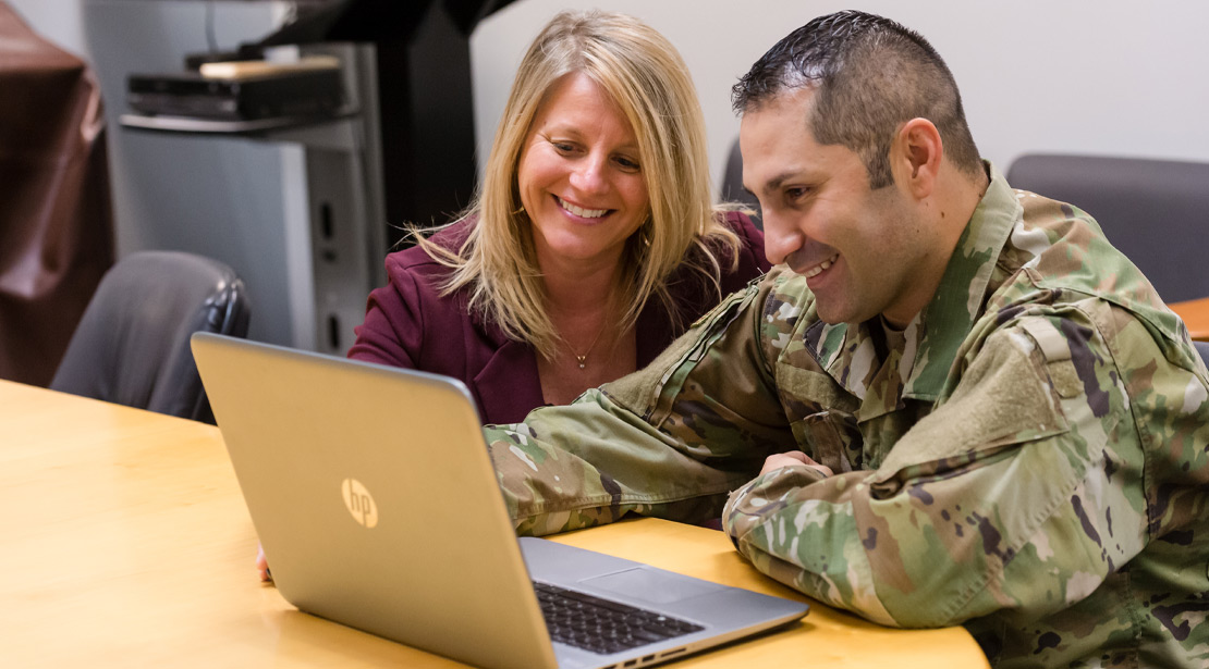Male student in fatigues at laptop with female teacher looking on and smiling. 