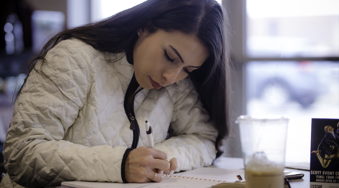 A young female student wearing a jacket and writing at a desk. 
