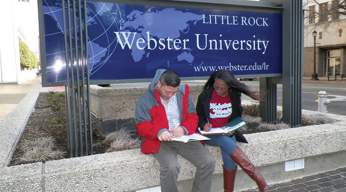 A male student and female student sitting in front of Little Rock Webster University sign, looking through papers. 