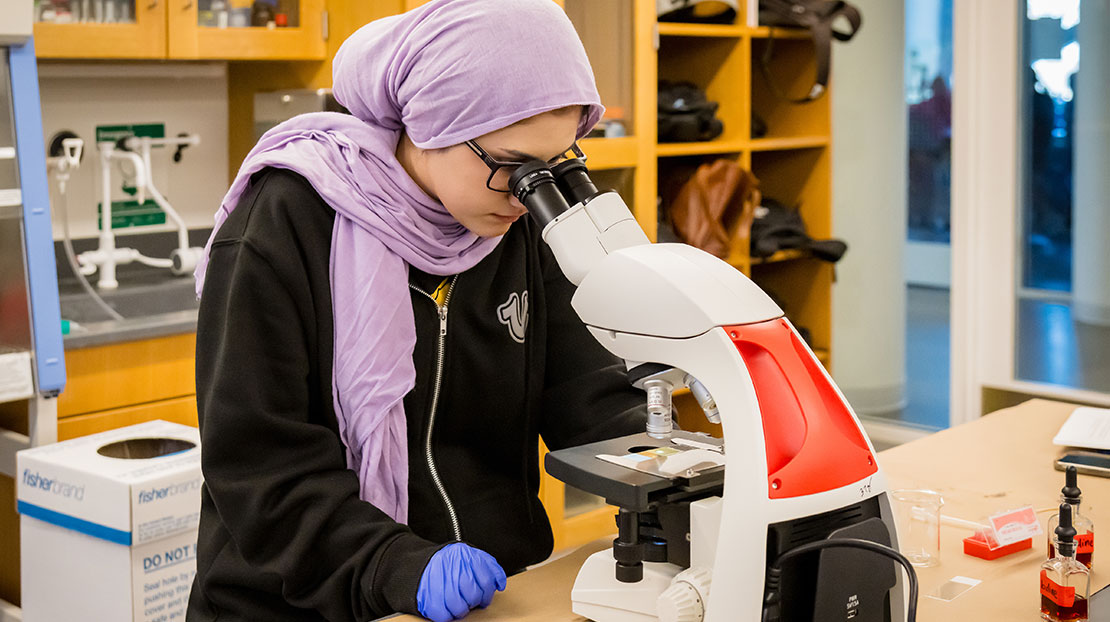 Student wearing lavender headscarf uses microscope