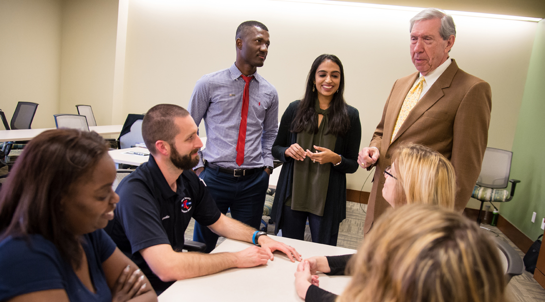 Group of Webster students, four sitting at a table, two standing near it with professor, talking.