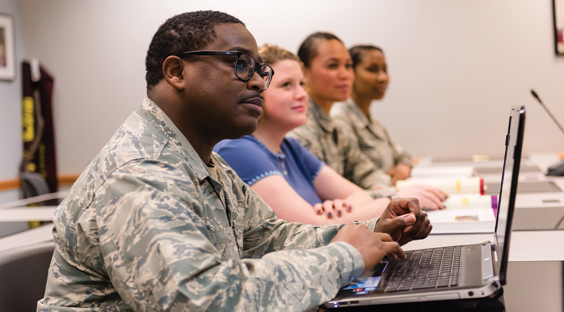 Four students sitting at table in classroom looking toward the front of the room and listening. 