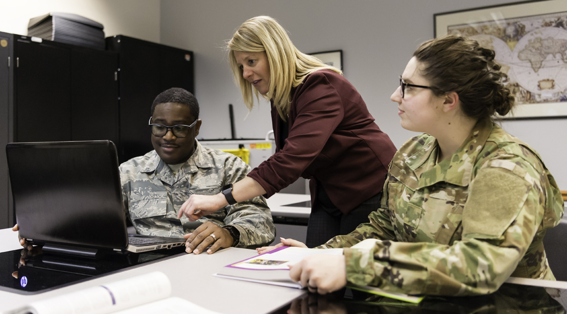 A female teacher points to a laptop screen while talking to a male student as a female student (both in fatigues) listens on. 