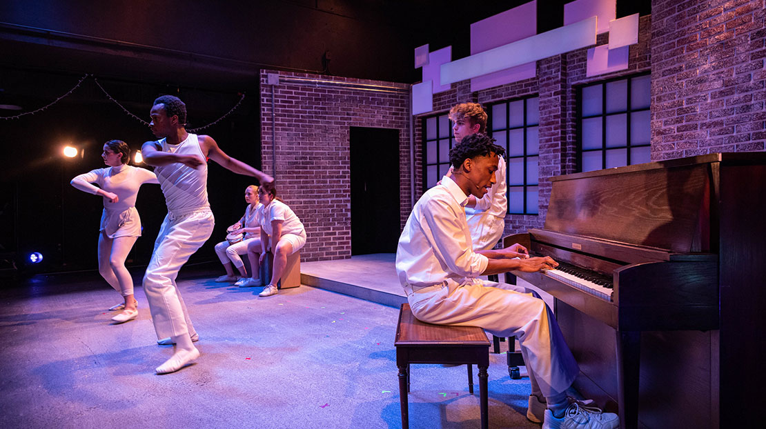 Person plays piano while two actors dance beside him, other actors sitting or standing on props, all wear white