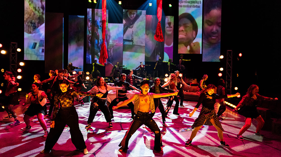 Backlit cast of characters dance energetically across stage with large image displays in background