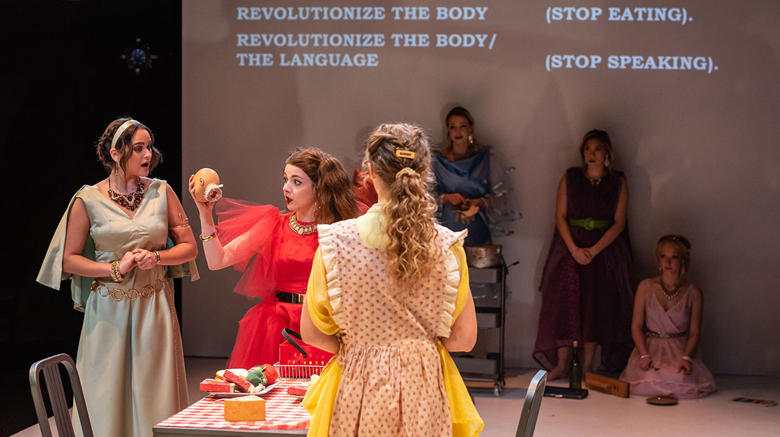 Three actresses around kitchen table, one pulls head off doll; words on screen in background reads Revolutionize the Body (Stop Eating), Revolutionize the Body/the Language (Stop Speaking)