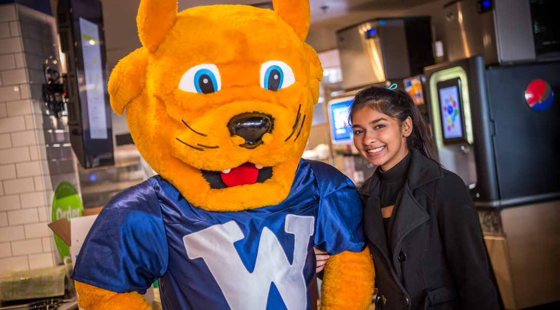 Gorlok mascot poses with a smiling female student with medium-dark skin tone wearing a black trenchcoat as they stand in the food area.