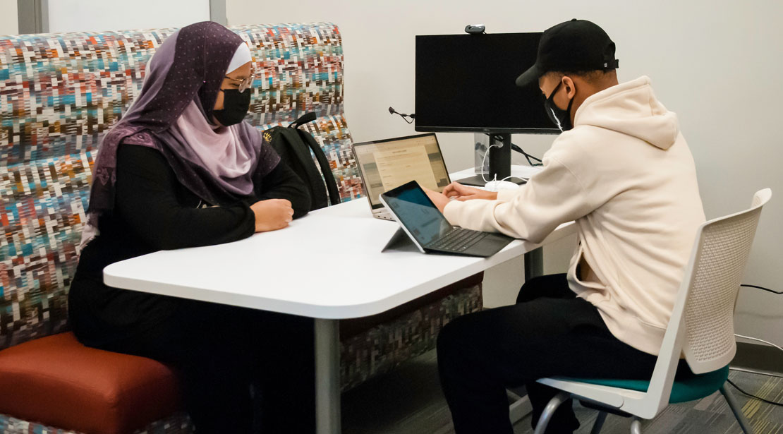 Students wearing face masks sit at a bench table with two laptops in front of a monitor with a web camera.