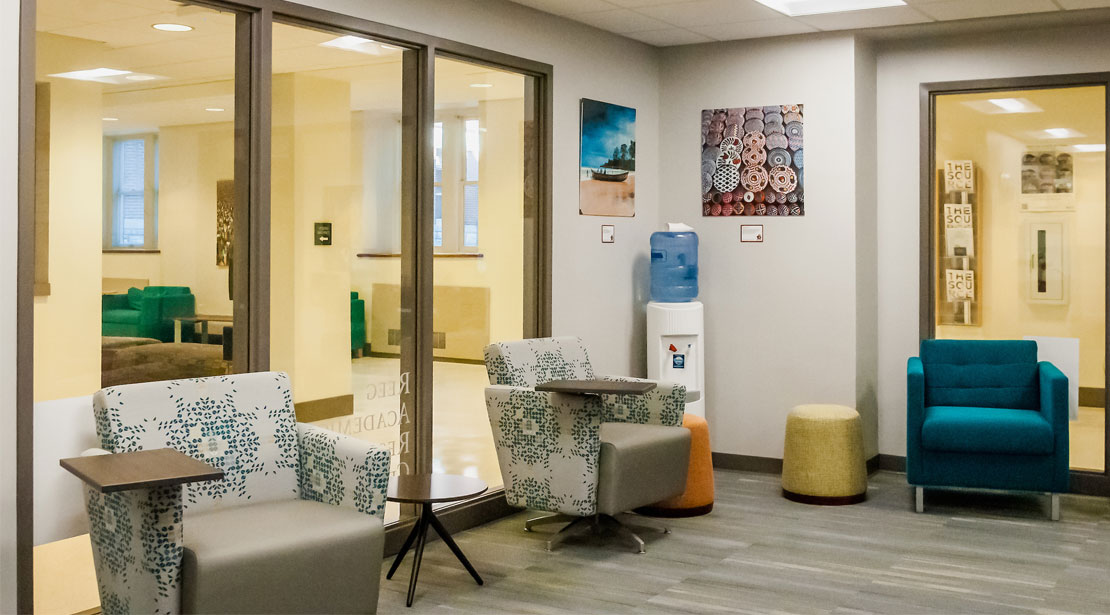 The lobby area of the Reeg Academic Resource Center has two comfortable turquoise chairs on the right side in front of a large window to the hallway, two small stools in yellow and orange in the middle in front of a water station, and two comfortable gray patterned chairs with a side desk attached.