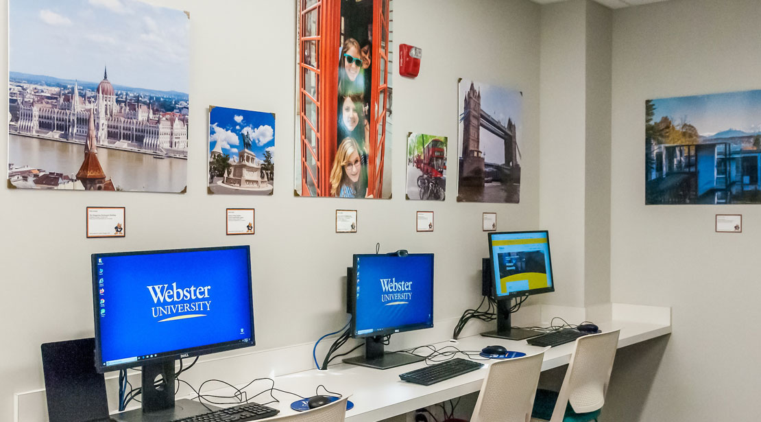 This is the hallway to the left of the welcome desk and shows a row of computer stations available for students.  There are chairs on wheels lining the long desks with computers." 