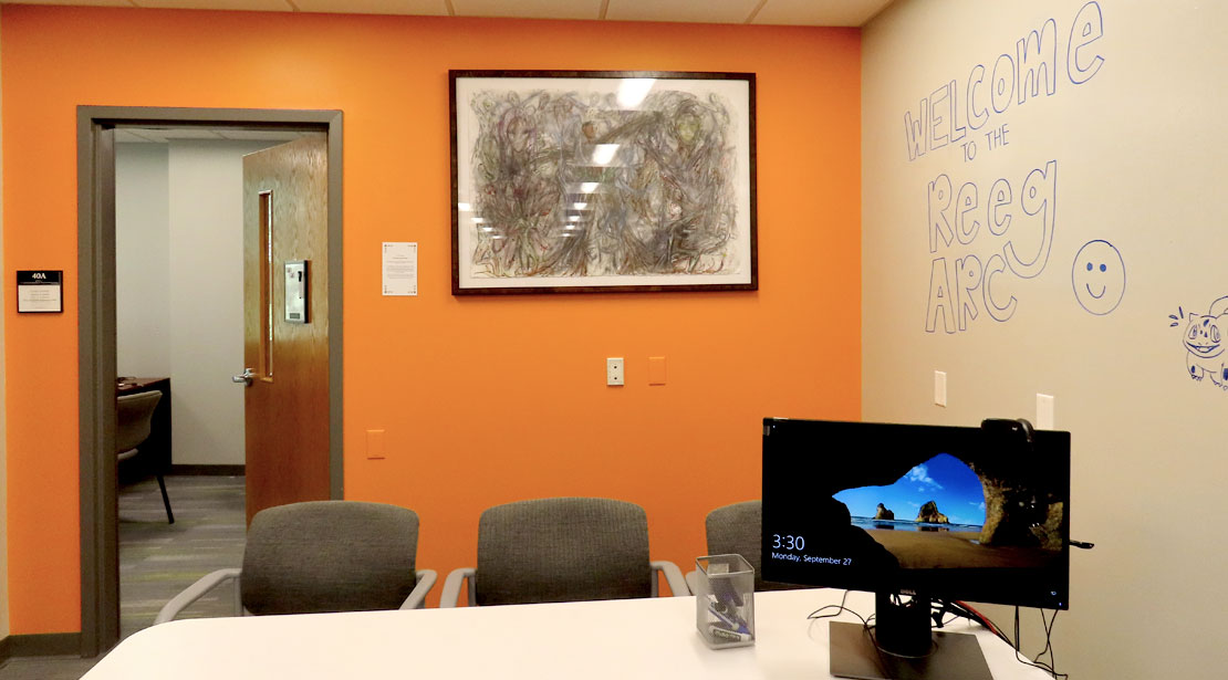 A room with a whiteboard wall has a table with three chairs on one side facing where a peer tutor would sit in front of a computer monitor to the right of the table.