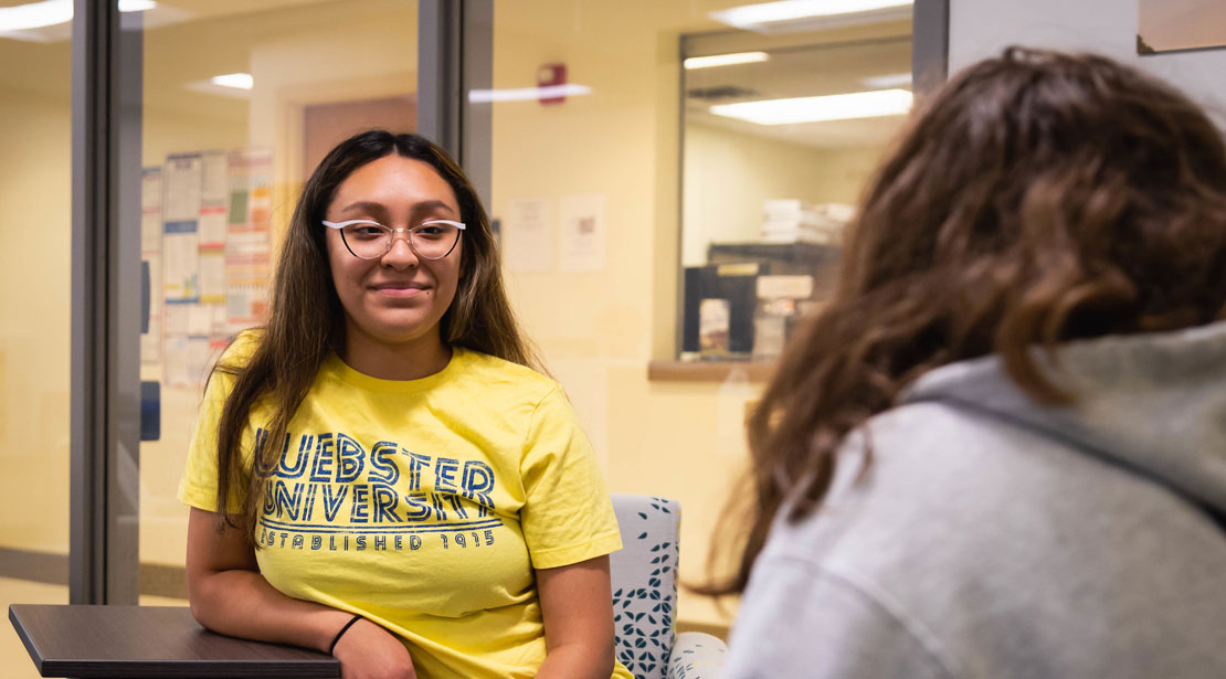 Two students are sitting in the lobby chairs; the young adult-aged female with long brown hair and glasses sits in a chair with an attached desk on the left and is smiling; the back of the head of the other student with brown hair is on the right.