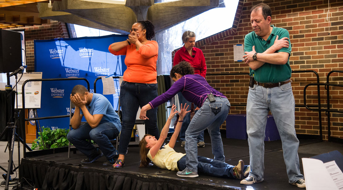 People from left to right: crouching with hands over eyes, covering mouth, on the floor with arms raised with one above them reaching down, in back of group holding self, and with arms crossed over upper arms.