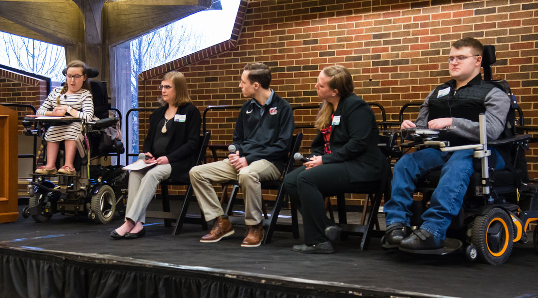 five people sitting on stage, two in wheelchairs