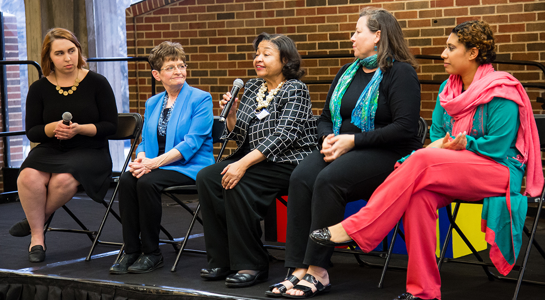 Five women sitting on stage, the center person with a microphone