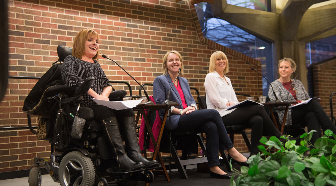 woman in wheelchair speaking, three women sit next to her on stage