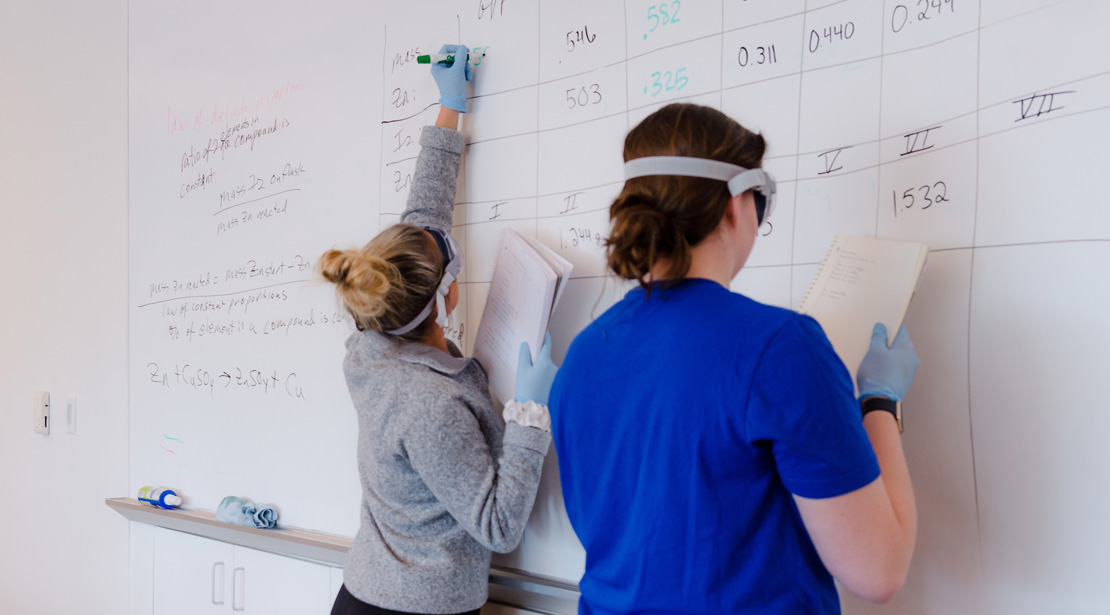 Two students writing experiment results on classroom whiteboard.