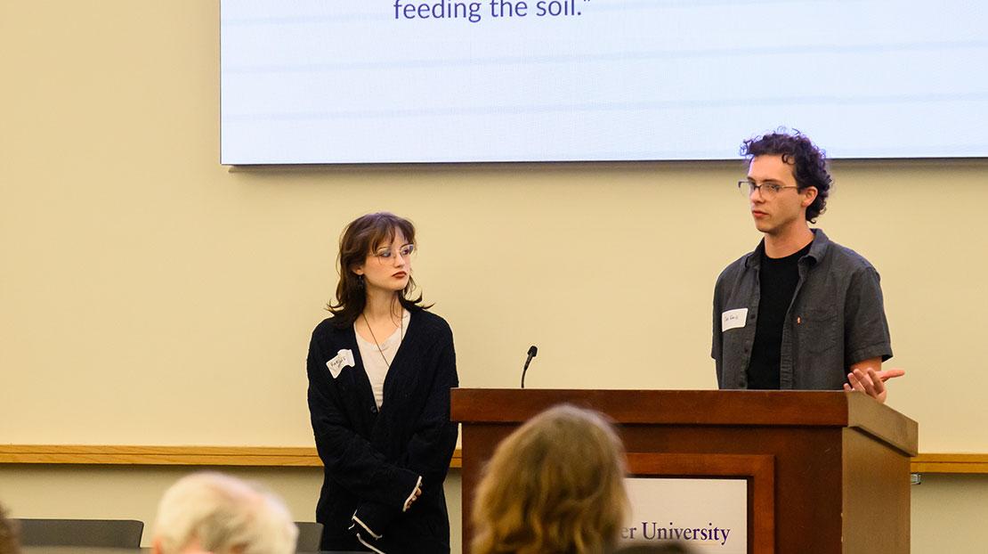 Reece Woodruff and Jack Farish stand underneath large screen to present about community gardens as sites for learning and change