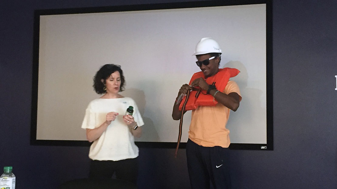 Professor holds vitamin jar while student wearing a hard hat adjusts life preserver over chest