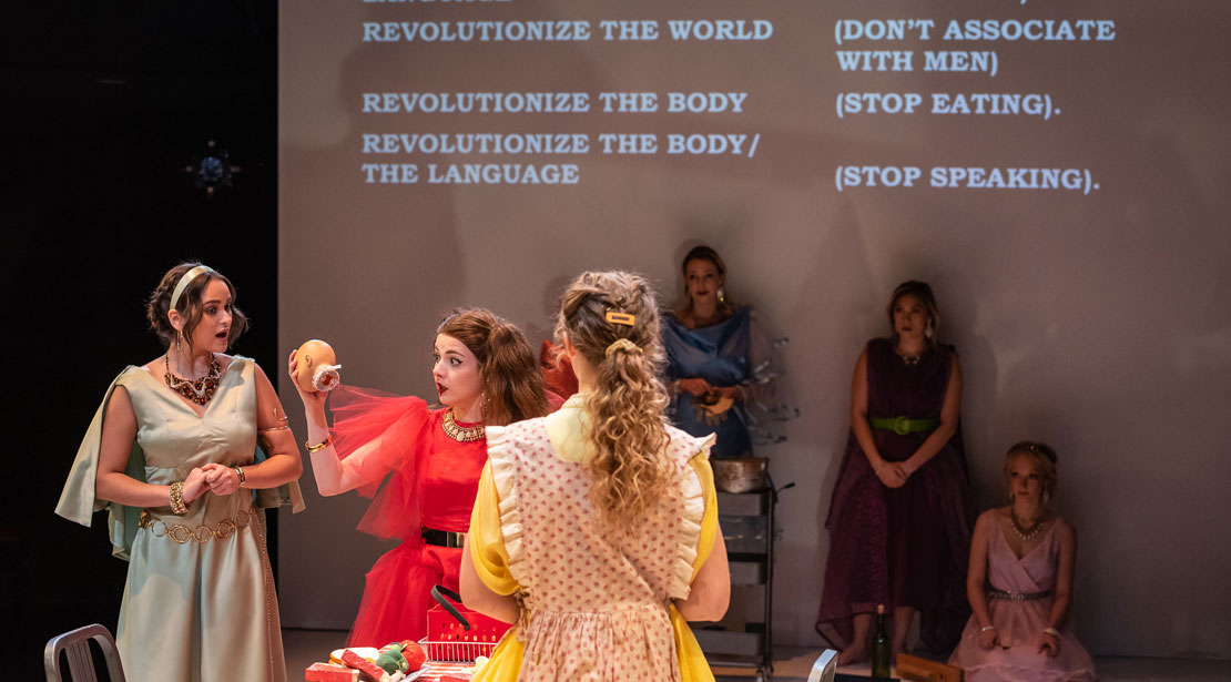Woman holding doll head in conversation with two others while a group of three are against a screen that reads, "Revolutionize the World. (Don't Associate with Men) Revolutionize the Body (Stop Eating) Revolutionize the Body/the Language (Stop Speaking)"
