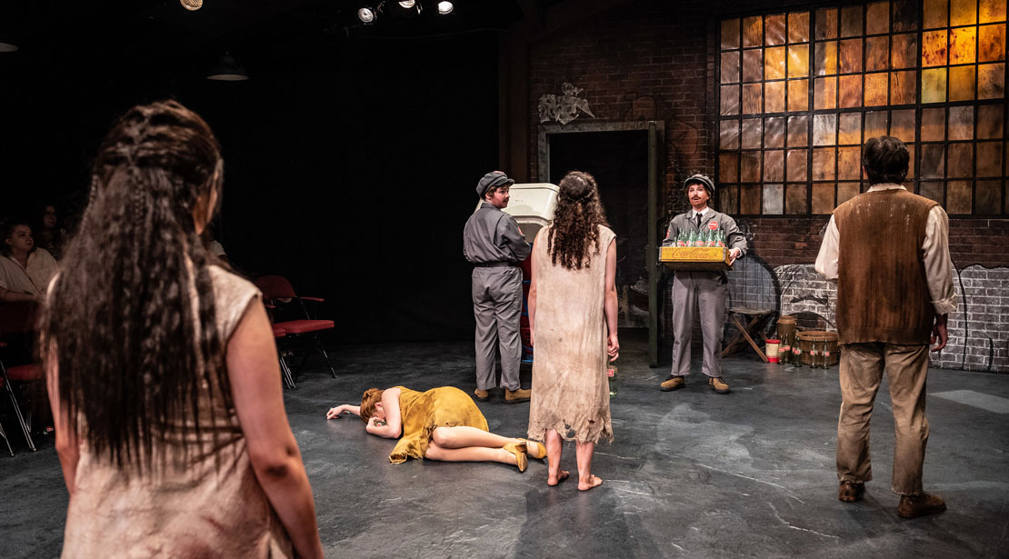 Two Coke delivery people stand in front of two women in dirty brown dresses and a man with a dark drown vest on; the woman in yellow is lying on the floor.