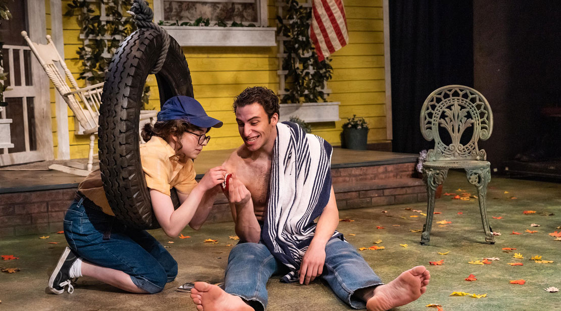 The actress sits on her knees leaning inside a tire swing talking with an actor sitting on the floor with legs straight out and feet angled outwards.