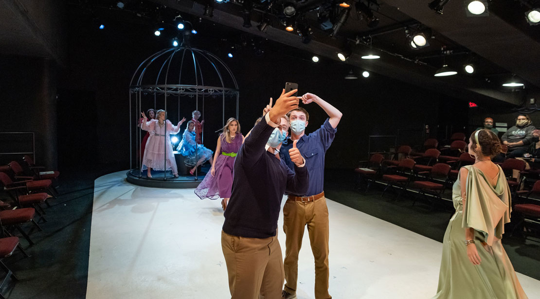 Two men wearing facemasks take selfies with women in a cage in the background while one woman catwalks the length of the stage next to them.