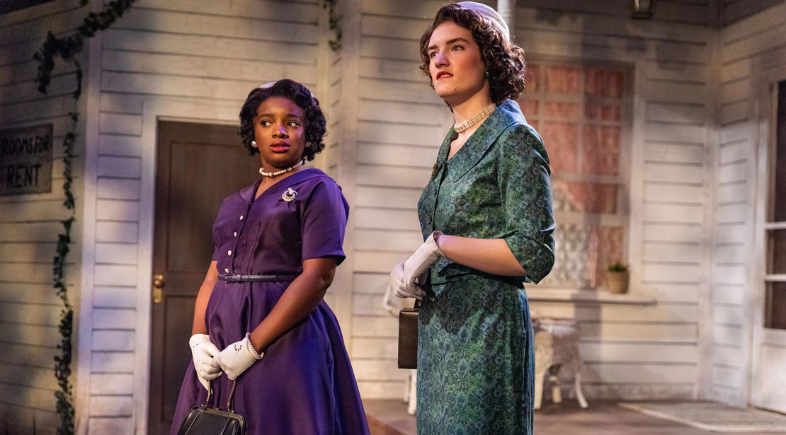 Two women in 1950s dresses wearing white gloves stand on the house porch looking on.