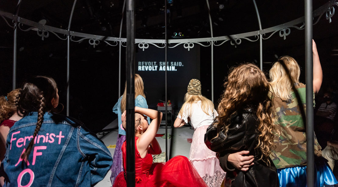 Seven women inside a cage looking across the theatre to a screen that reads, "Revolt. She Said. Revolt Again."