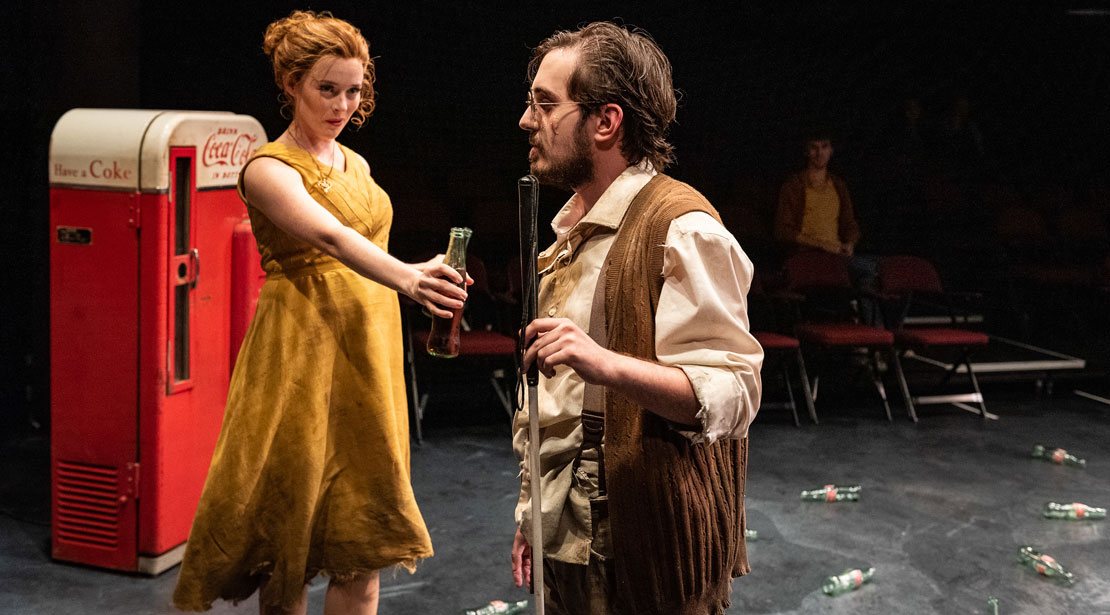 A woman in a yellow dress with red hair holds out a full Coke bottle to a blind man.