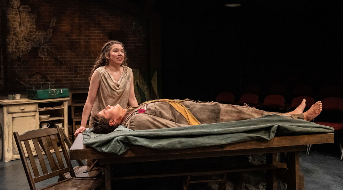 A woman with brown hair in a dirty and tattered brown dress stands over a table with a still man laying on it wearing dirty, brown clothing.