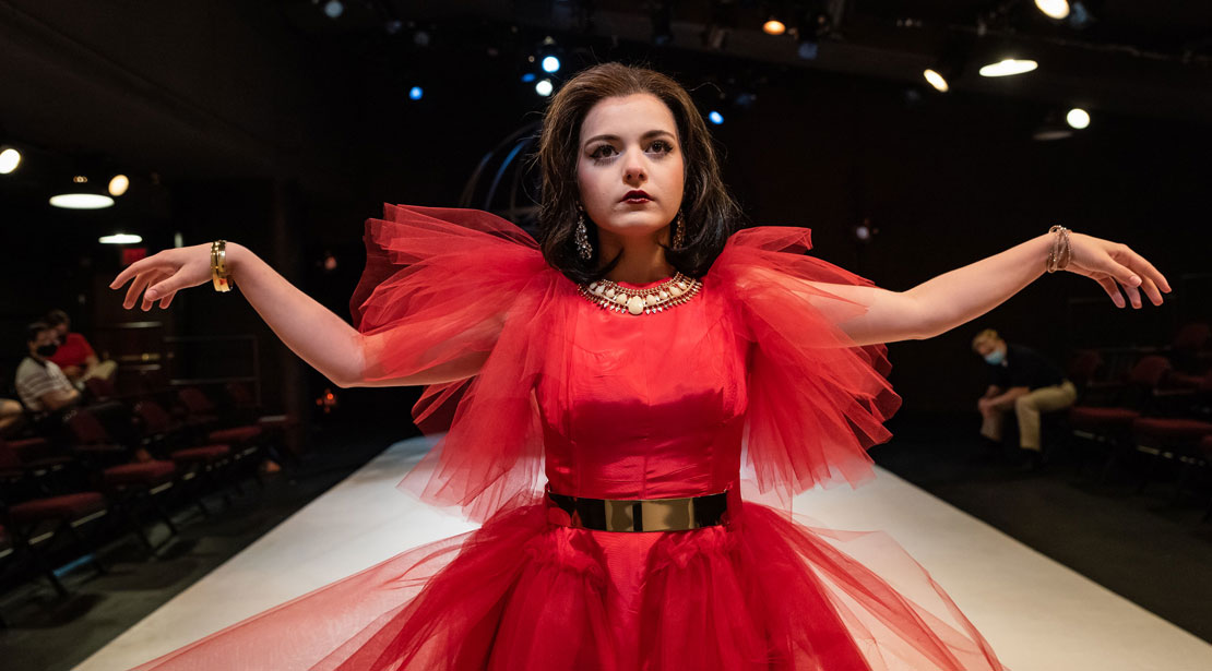 Actress with brown shoulder-length hair wearing a red dress with crinoline sleeves poses at the end of the runway with arms held out to sides with bent elbows and wrists.