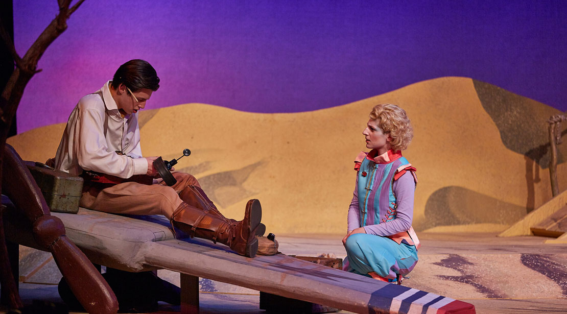 The pilot sits on his plane wing while the actor in blue, purple and red kneels in front of him
