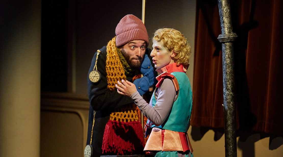 An actor with a knit cap and a stopwatch over their shoulder talks conspiratorily with the actor with the blue, purple, and red outfit