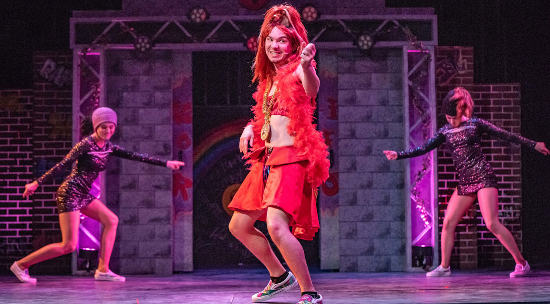 An actor wearing a red halter top, skirt, and boa with long red hair dancing with two actors in black short dresses dancing in the background