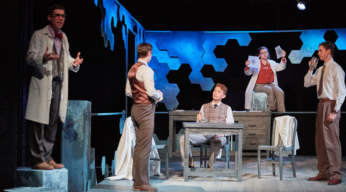 A laboratory scene with five actors, one in the front talking to the audience, one at a desk talking to two others, and the fifth looking at photos in the back