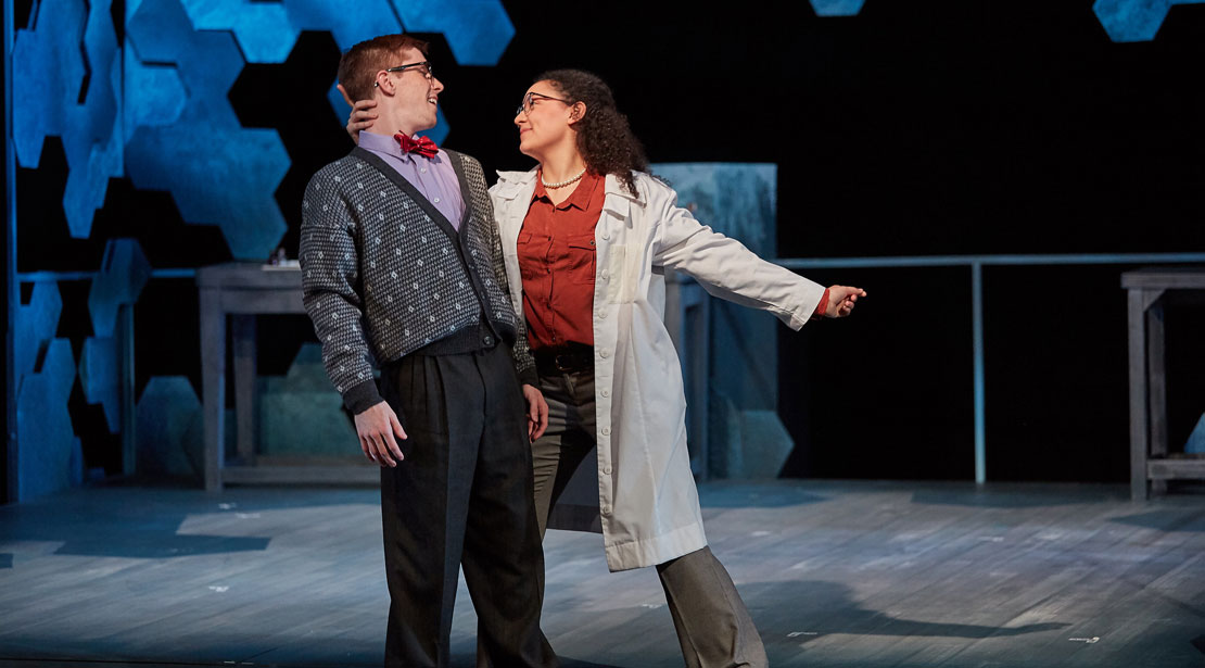 A female scientist holds another actor's head while dipping him backward