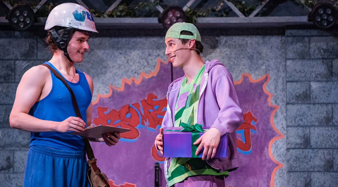 An actor wearing a blue tank top and shorts with a bike helmet on talks to an actor wearing blue and purple clothes holding a package