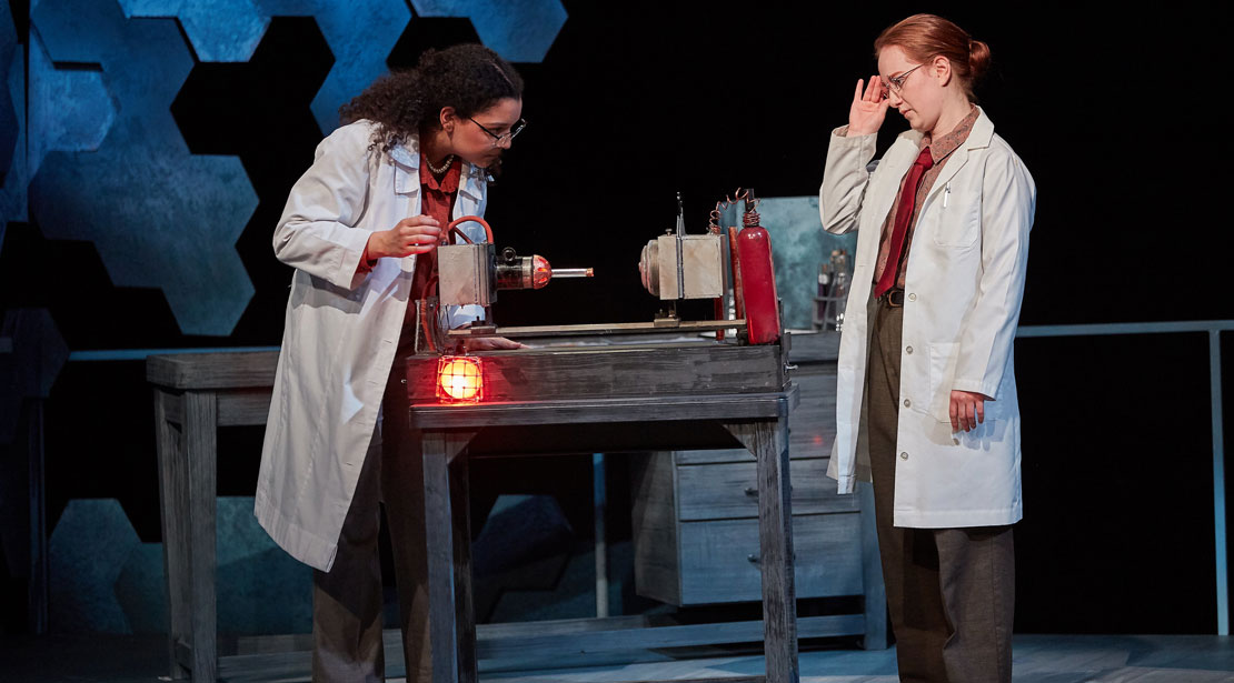 Two actresses in lab coats examining device