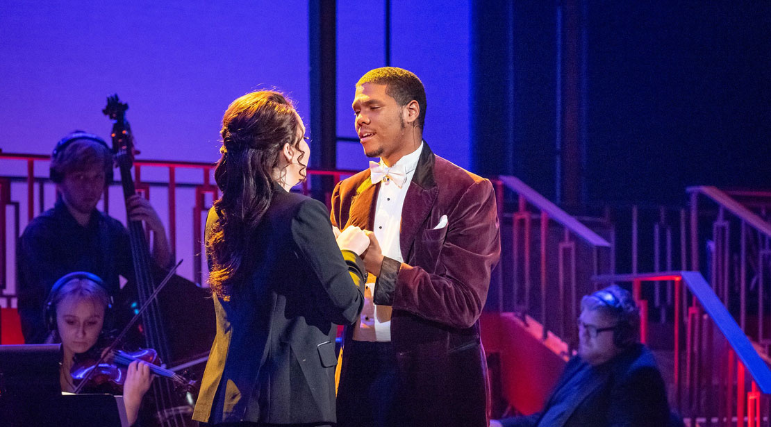 Two actors sing together in front of an orchestral group with a purple-lighted background