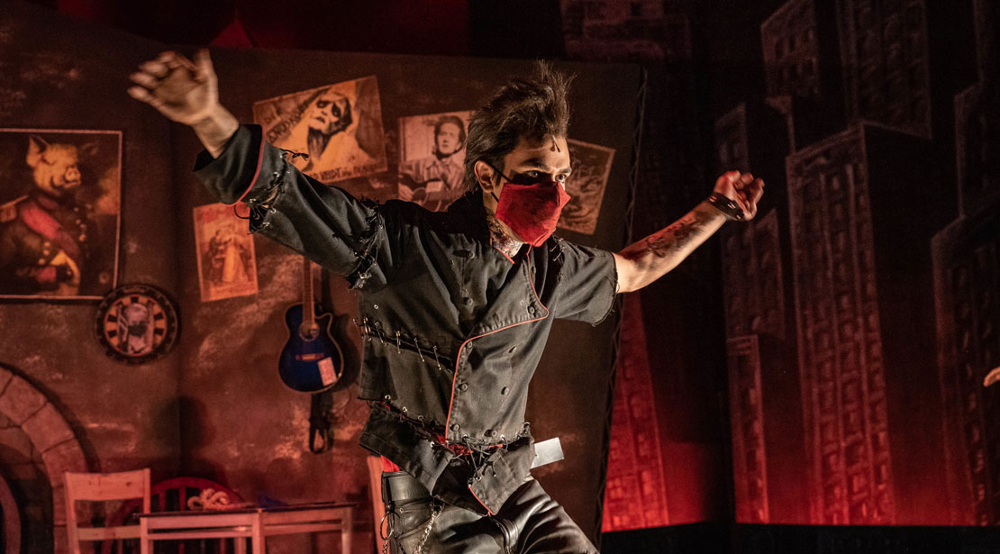 An actor wearing a black shirt and pants with rips that are closed with exposed safety pins, wearing a red mask with arms thrown wide