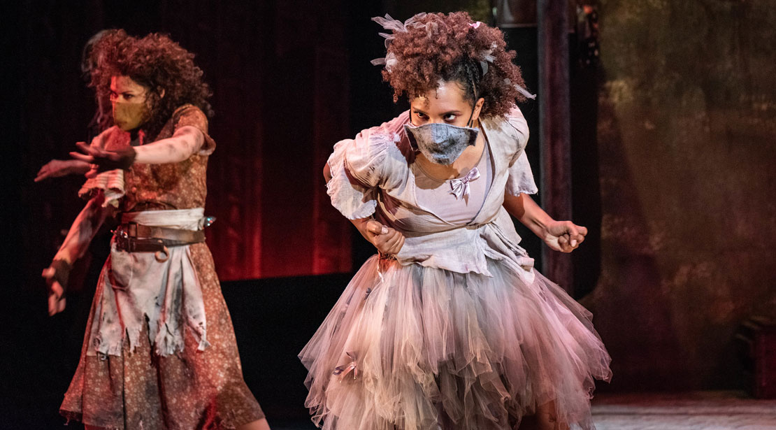 Two masked actors in ripped dresses stand in aggressive poses with clenched fists