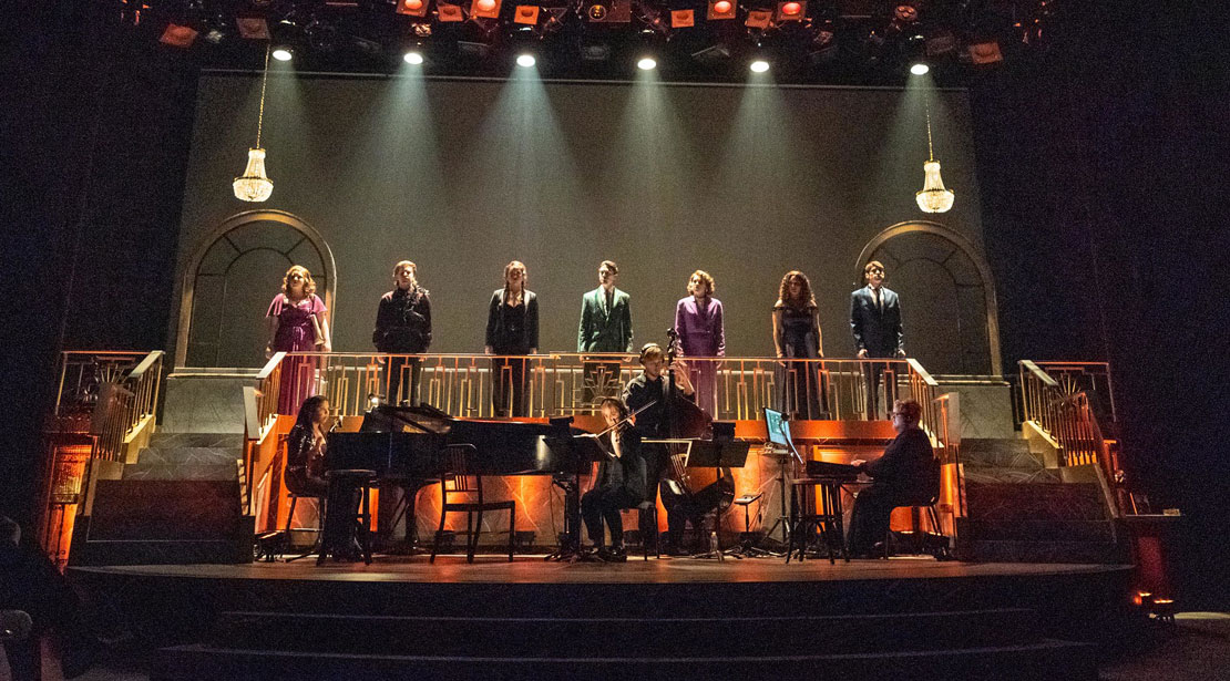 On a backlit stage, seven actors stand on the top section of the stage behind the band.