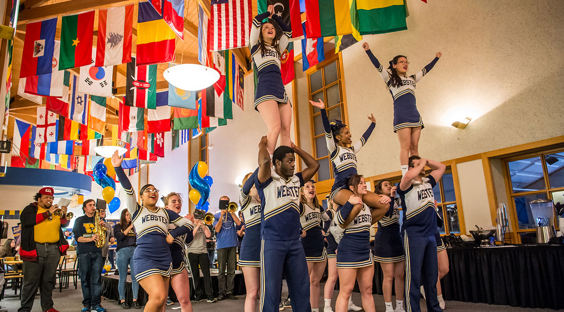 Webster cheerleaders perform several lifts inside a room with dozens of national flags from different nations suspended from the ceiling.