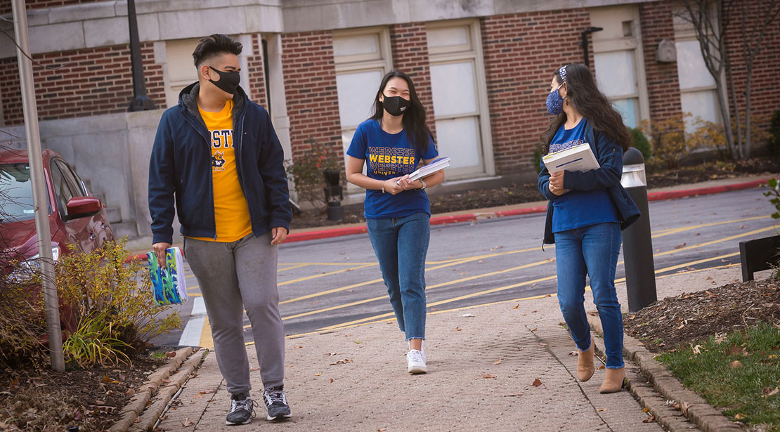 Three students walked down a brick sidewalk together. They are wearing masks and practicing social distancing.